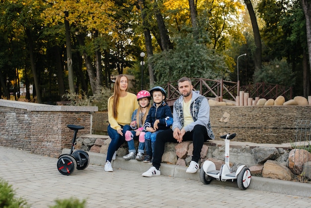 Une grande famille heureuse monte des Segways et des scooters électriques dans le parc par une chaude journée d'automne au coucher du soleil