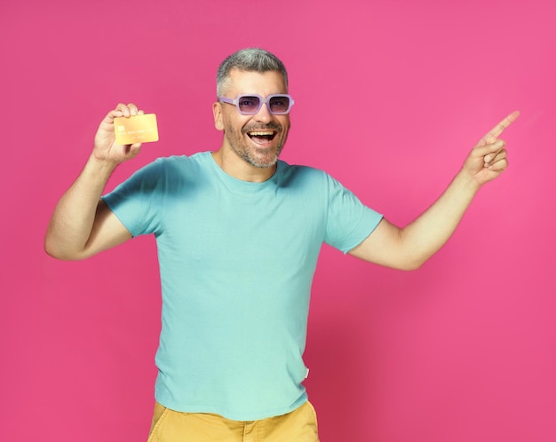 Photo grande offre bel homme tenir une carte de crédit de débit à la main pointant sur le côté portant un t-shirt bleu et des lunettes isolés sur fond rose homme avec carte bancaire en main concept bancaire financier