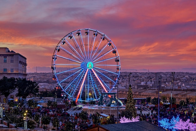 Grande roue colorée contre le ciel rose au coucher du soleil et le paysage urbain de Malte. Marché de Noël à La Valette Malte vue aérienne, flou de mouvement, mise au point sélective