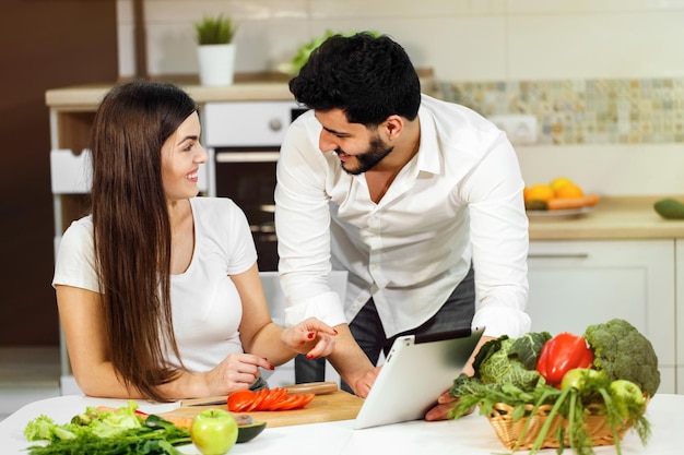 Grande soirée familiale jolie femme aux cheveux noirs coupant la tomate beau grand homme montrant des photos sur une tablette prise de vue en intérieur dans une belle cuisine blanche concept d'une alimentation saine et d'un beau corps