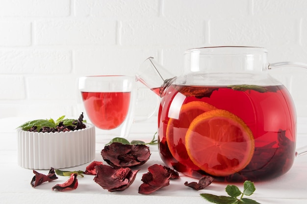 Grande théière en verre avec fruits et tisane thé rouge sur fond de mur de briques blanches thé vitaminé