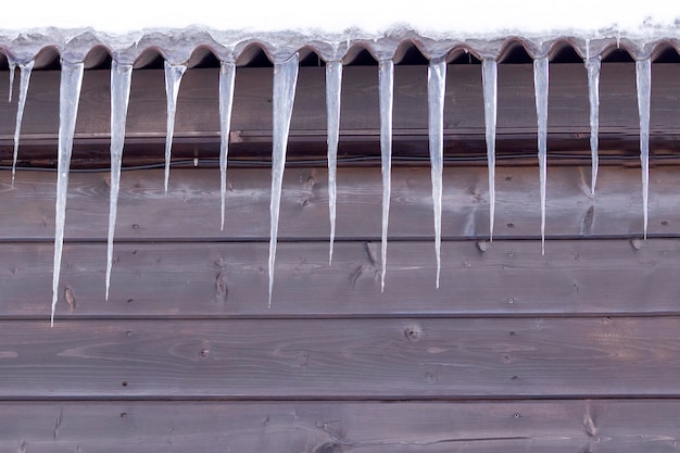 De grands glaçons pendent du toit enneigé d'une maison en bois Fond d'hiver