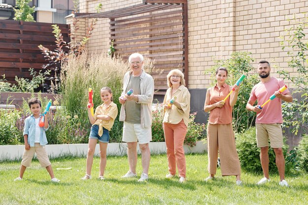 Grands membres de la famille debout sur une pelouse verte s'amusant à poser avec des pistolets à eau