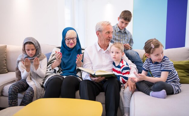 grands-parents de la famille musulmane moderne avec petits-enfants lisant le Coran et priant ensemble sur le canapé avant le dîner de l'iftar lors d'une fête du ramadan à la maison