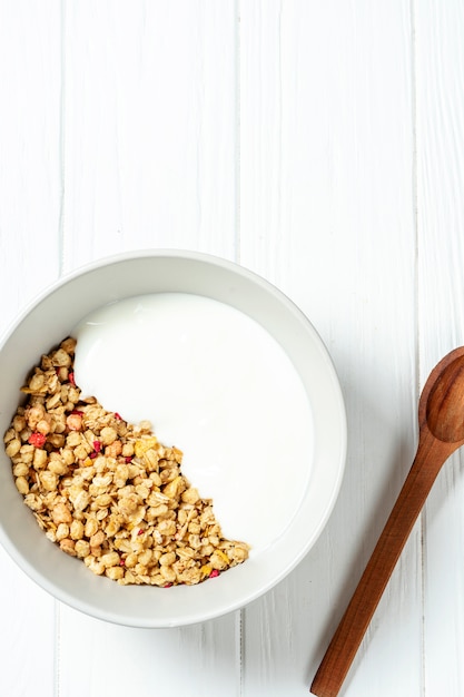 Granola de blé blanc avec du yogourt faible en gras dans un bol blanc dans une composition avec une cuillère sur fond de bois blanc.