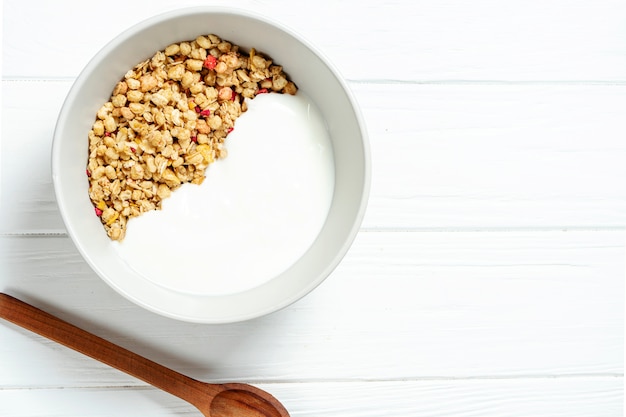 Granola de blé blanc avec du yogourt faible en gras dans un bol blanc dans une composition avec une cuillère sur fond de bois blanc.