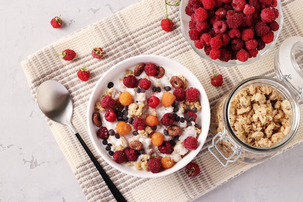 Granola croustillant avec yaourt naturel, framboises rouges et jaunes fraîches, chocolat et noix dans un bol blanc sur fond gris. Vue d'en-haut