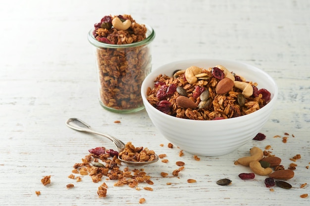 Granola fait maison dans un bocal en verre avec du yaourt grec ou du lait et des noix de cajou citrouille aux amandes avec des graines de canneberges séchées sur fond de table en bois rustique blanc Petit-déjeuner ou collation énergétique sain Vue de dessus