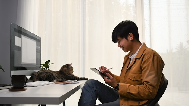 Graphiste décontracté de jeune homme assis avec son chat au bureau à domicile et travaillant avec une tablette numérique.