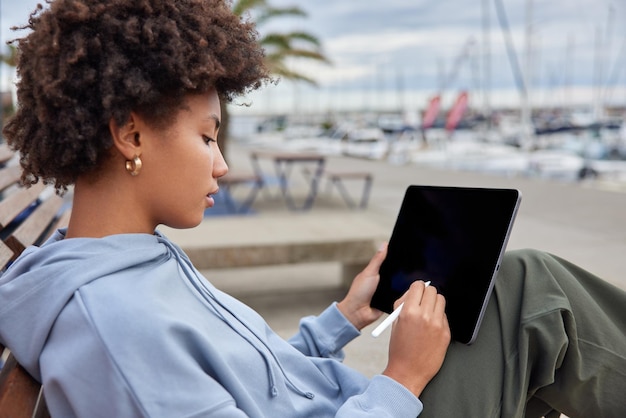 Un graphiste expérimenté dessine des croquis sur une tablette numérique utilise un stylet crée la conception du logo porte un sweat à capuche décontracté pose de côté à la caméra a une occupation créative passe du temps libre dans le port.