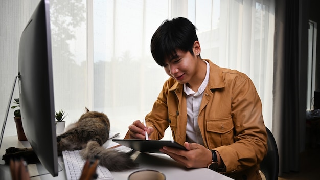 Graphiste souriant de jeune homme travaillant en ligne avec une tablette numérique et son chat allongé devant lui.