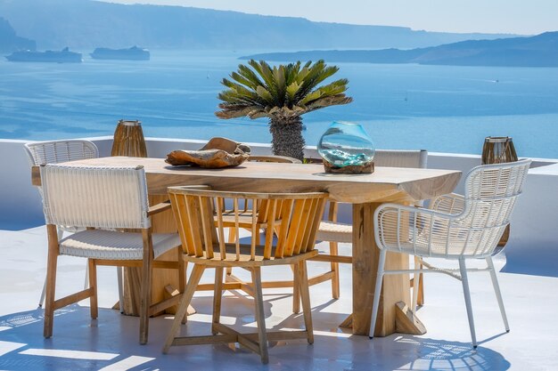 Grèce. Santorin. L'île de Thira. Table et chaises en bois sur une terrasse ensoleillée. Deux bateaux de croisière dans le port