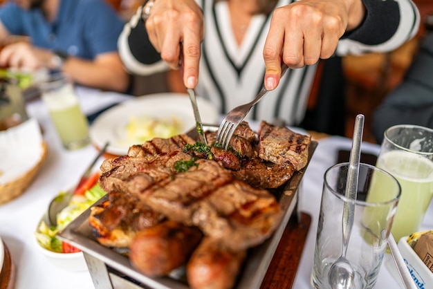 Photo un gril plein de viande de bœuf, de poulet et de chorizo. les mains d'une femme coupant de la viande.