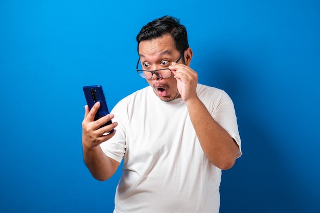 Un gros asiatique portant un t-shirt blanc a l'air surpris des bonnes nouvelles qu'il a reçues de son smartphone. Les hommes montrent des mouvements choqués avec des yeux exorbités tout en baissant leurs lunettes sur un smartphone