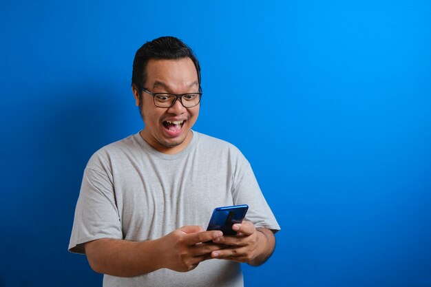 Un gros asiatique portant un T-shirt gris a l'air heureux des bonnes nouvelles qu'il a reçues de son smartphone. Les hommes montrent des gestes de victoire. fond bleu