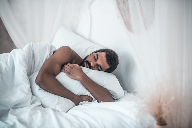 Gros dormeur. Jeune adulte à la peau sombre homme séduisant dormir étreignant oreiller blanc dans la chambre lumineuse