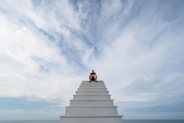 Un gros homme asiatique se tient sur un escalier en bois avec un fond de ciel bleu et un horizon sans fin et l'océan