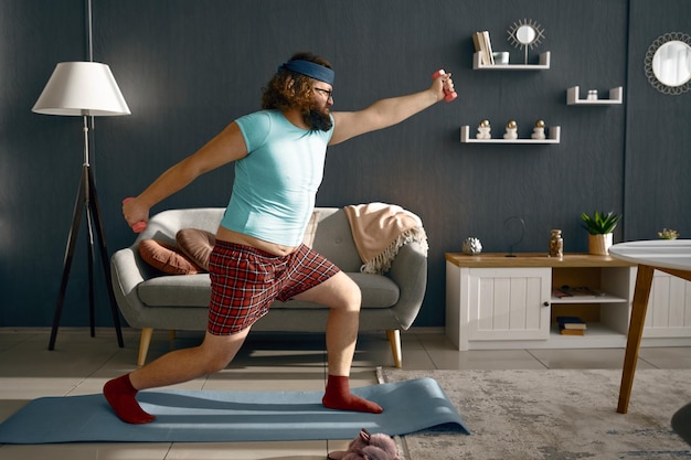 Un gros homme drôle fait de l'exercice le matin avec des haltères à la maison. Sport, mode de vie sain et perte de poids