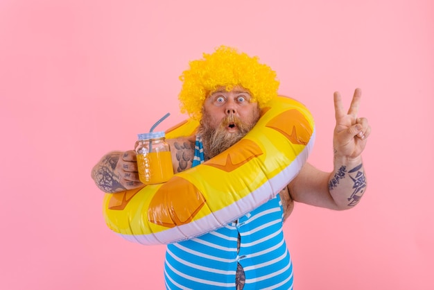 Un gros homme étonné avec une perruque dans la tête est prêt à nager avec une bouée de sauvetage en forme de beignet