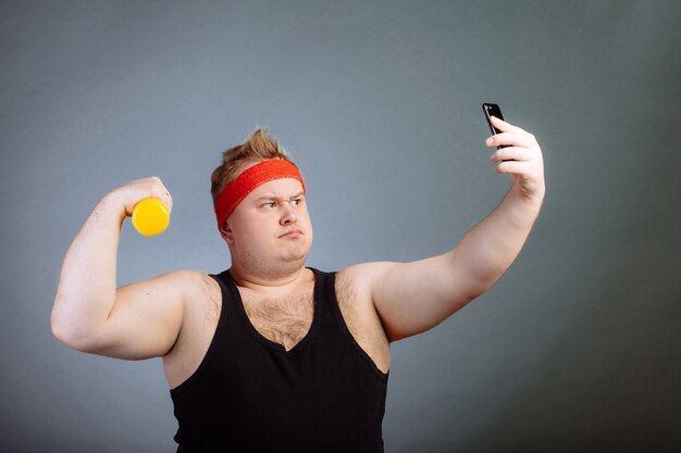 Gros homme avec gros ventre, tenant des haltères, faisant selfie sur mur gris