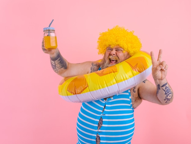 Un gros homme heureux avec une perruque dans la tête est prêt à nager avec une bouée de sauvetage en forme de beignet