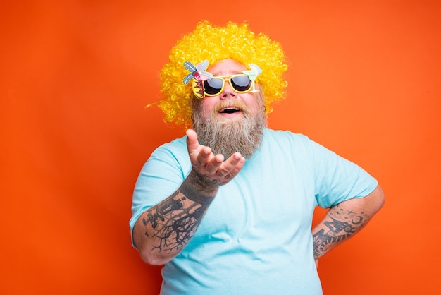 Gros homme heureux avec des tatouages de barbe et des lunettes de soleil s'amuse avec la perruque jaune