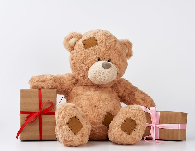 Gros ours en peluche beige avec des correctifs, une pile de cadeaux dans des boîtes enveloppées dans du papier brun, noué avec un ruban de soie sur un fond blanc