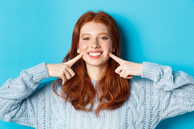 Gros plan d'une adolescente joyeuse aux cheveux rouges et taches de rousseur, piquer les joues, montrant des fossettes et souriant avec des dents blanches, debout sur fond bleu.