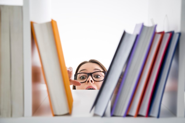 Gros plan d'une adolescente portant des lunettes et atteignant un livre orange debout sur une étagère haute de la bibliothèque