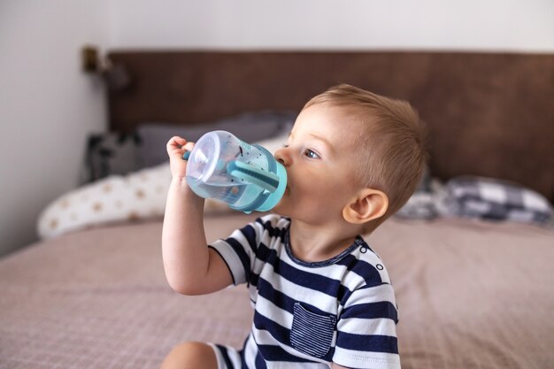 Gros plan d'adorable petite blonde assise sur le lit dans la chambre et de l'eau potable de sa bouteille.