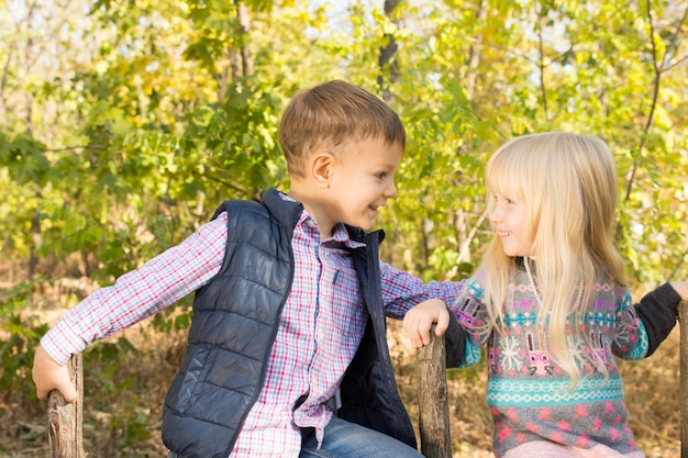 Gros Plan D'adorables Petits Enfants Blancs En Tenue De Mode D'automne Souriant Les Uns Aux Autres Dans Le Parc Avec Des Arbres Verts à L'arrière-plan.