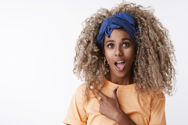 Gros plan amusé impertinent et surpris femme afro-américaine aux cheveux bouclés blonds avec bandeau bleu pointant le coin supérieur gauche espace de copie vierge haletant fasciné et impressionné fond blanc