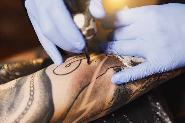 Gros plan d'un artiste tatoueur professionnel faisant un tatouage sur le bras d'un jeune homme par machine avec de l'encre noire