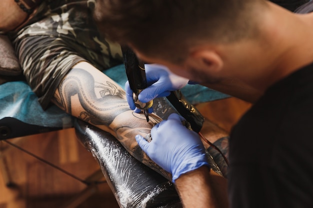 Gros plan d'un artiste tatoueur professionnel faisant un tatouage sur le bras d'un jeune homme par machine avec de l'encre noire