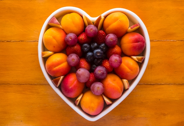 Gros Plan D'une Assiette Appétissante De Fruits Frais Mûrs De Saison Dans Une Assiette Blanche En Forme De Coeur - Alimentation Saine Avec Abricots, Raisins, Fraises, Myrtilles Et Pommes - Fond En Bois