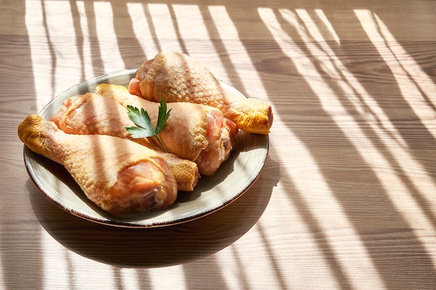 Gros plan d'une assiette avec des cuisses de poulet crues sur une table en bois à côté d'une fenêtre qui laisse entrer les rayons du soleil entre les lumières et les ombres