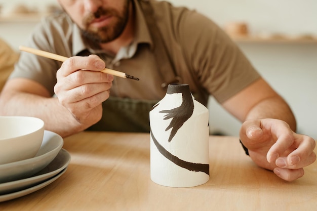 Gros plan aux tons chauds d'un homme méconnaissable décorant des céramiques dans un atelier de poterie, espace pour copie