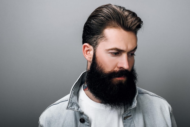 Gros plan d'un beau hipster à la mode jeune homme de race blanche avec une barbe épaisse portant une veste en jean à la mode ayant un regard confiant et sérieux Barber man posant contre le mur gris du studio