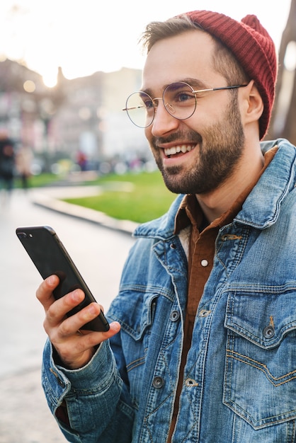 Gros plan d'un beau jeune homme barbu élégant marchant à l'extérieur dans la rue, à l'aide d'un téléphone portable