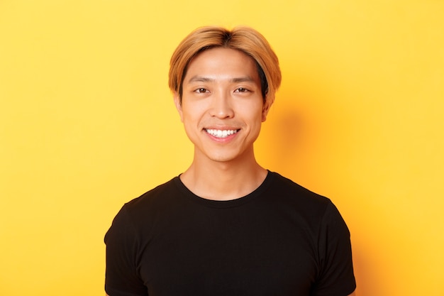 Gros plan d'un beau mec asiatique blond en t-shirt noir, souriant heureux, debout sur un mur jaune.