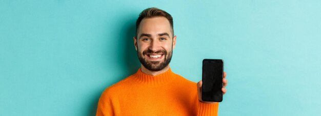 Gros plan d'un beau mec barbu en pull orange montrant l'écran du smartphone et souriant montrant le bal