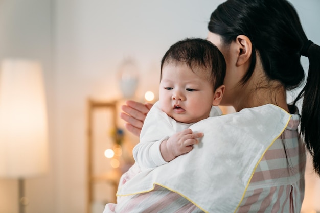 gros plan bébé asiatique innocent appuyé sur l'épaule de sa mère avec un chiffon regarde loin dans l'espace pendant que sa mère rote en tapotant sur son dos à la maison.
