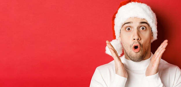 Gros plan d'un bel homme étonné en bonnet de Noel et chandail blanc levant les mains et l'air impressionné st...