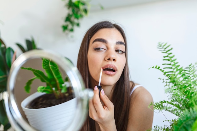 Gros plan belle femme appliquant du brillant à lèvres devant un petit miroir avec des plantes autour