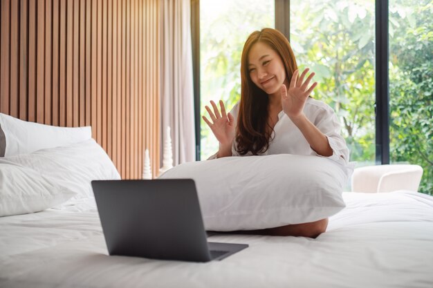 Gros plan d'une belle femme asiatique agitant les mains tout en utilisant un ordinateur portable pour un appel vidéo tout en étant assis sur un lit à la maison