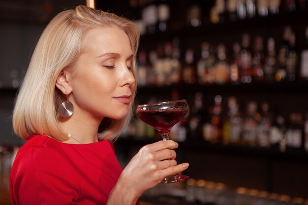 Gros plan d'une belle femme blonde souriante, les yeux fermés, prendre un verre au bar le soir