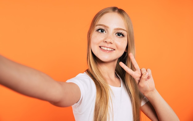 Gros plan d'une belle fille blonde élégante et branchée en t-shirt blanc pendant qu'elle fait une photo de selfie sur son téléphone intelligent isolé en studio