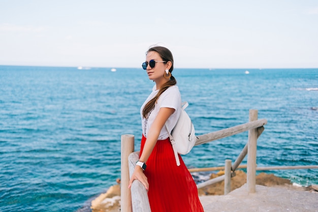 Gros plan d'une belle fille à lunettes rondes et une robe rouge près d'une balustrade en bois sur un rocher surplombant la mer