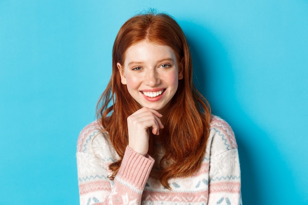 Gros plan sur une belle fille rousse souriante, ayant une conversation et regardant la caméra intéressée, debout sur fond bleu.