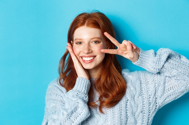 Gros plan sur une belle fille souriante aux cheveux rouges, montrant le signe de la paix kawaii et regardant la caméra, debout sur fond bleu.